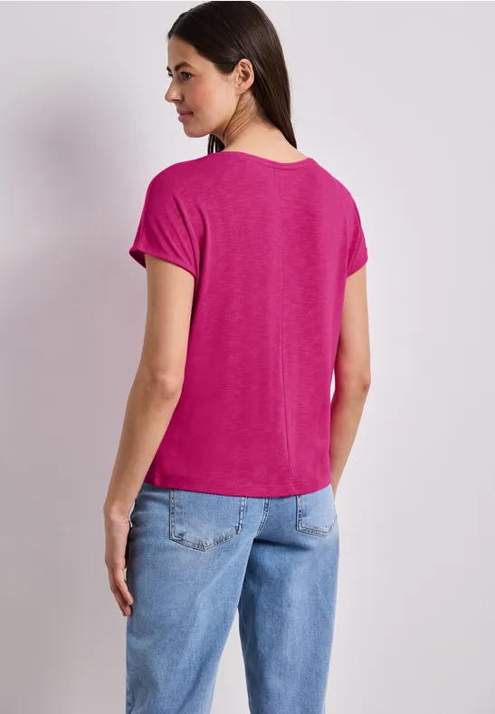 Frau von hinten, trägt pinkes T-Shirt und blaue Jeans, steht vor hellem Hintergrund.