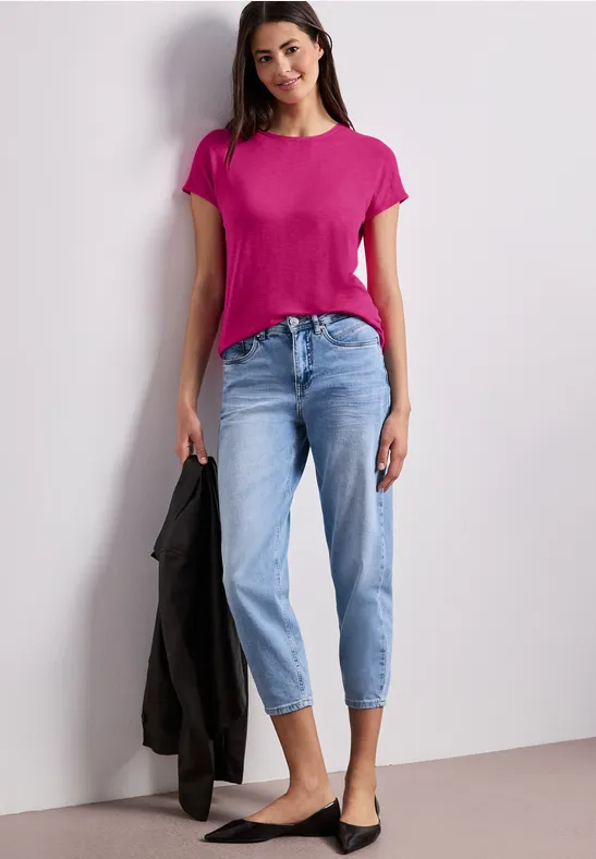 Frau in pinkem T-Shirt und hellblauen Jeans, hält schwarze Jacke, trägt schwarze Ballerinas vor hellem Hintergrund.