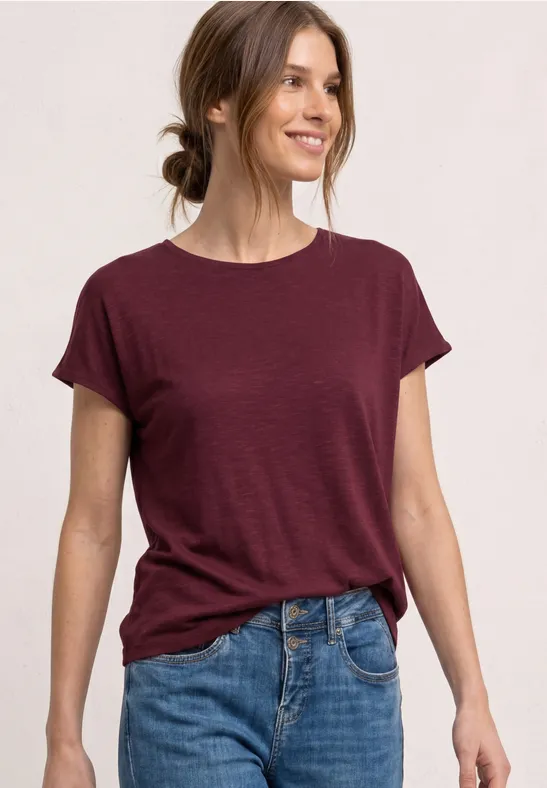 Een vrouw draagt een wijnrood T-shirt en blauwe jeans voor een lichte achtergrond.