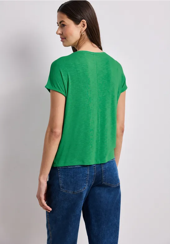 Frau trägt grünes T-Shirt und blaue Jeans, steht mit dem Rücken zur Kamera vor hellem Hintergrund.