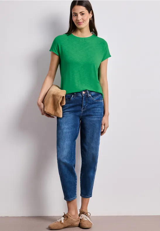 Frau in grünem T-Shirt und blauen Jeans steht vor Hintergrund, hält Jacke in der Hand.