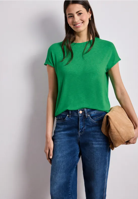 Eine Person trägt ein grünes T-Shirt, blaue Jeans und hält eine beige Tasche in der Hand.