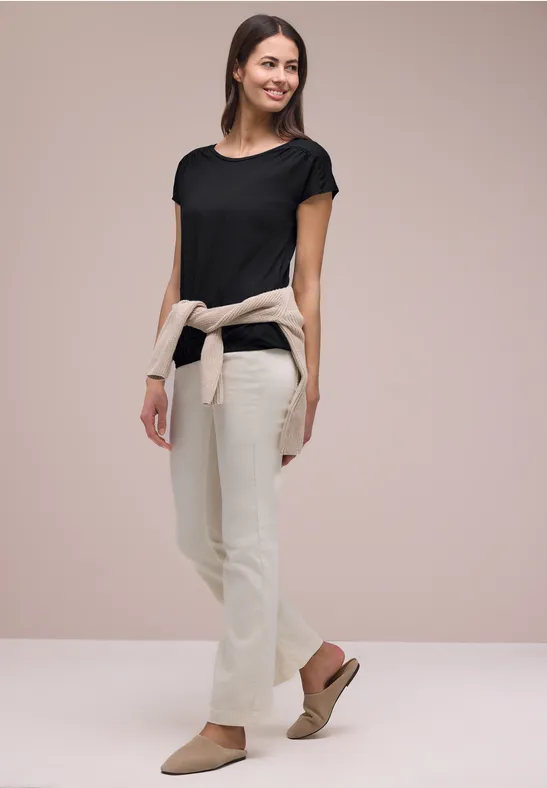 Vrouw in zwart T-shirt, lichte broek, trui om de taille geknoopt en sandalen.