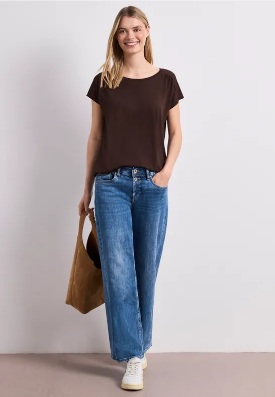 Vrouw in bruin T-shirt, wijde spijkerbroek, witte sneakers met bruine tas tegen lichte achtergrond.