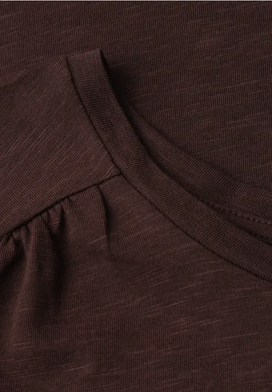 Close-up van een donkerbruin T-shirt met ronde hals en subtiele plooien bij de schouders.