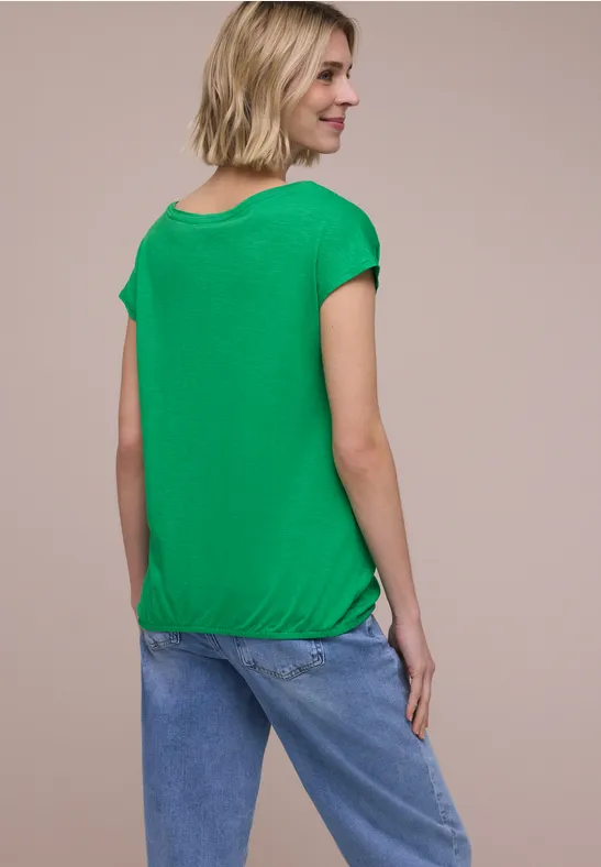 Vrouw van achteren in groene top en blauwe jeans tegen een neutrale achtergrond.