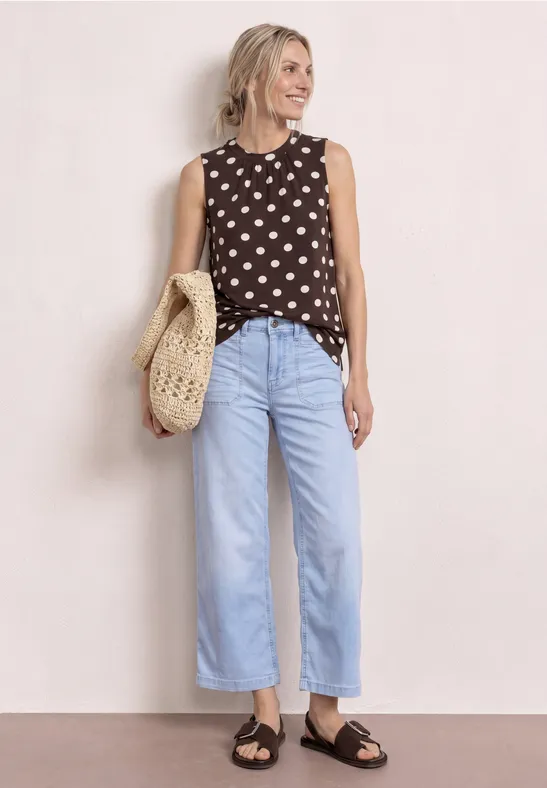 Eine Person trägt ein ärmelloses gepunktetes Top, weite hellblaue Jeans, schwarze Sandalen und eine gehäkelte Tasche.
