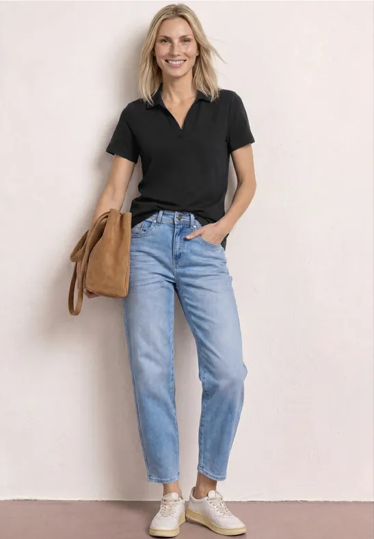 Vrouw in zwart poloshirt, jeans en witte sneakers, staat voor een lichte muur met een beige schoudertas.