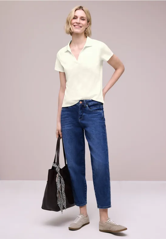 Vrouw in wit poloshirt, blauwe jeans en beige loafers met zwarte tas en sjaal voor grijze achtergrond.