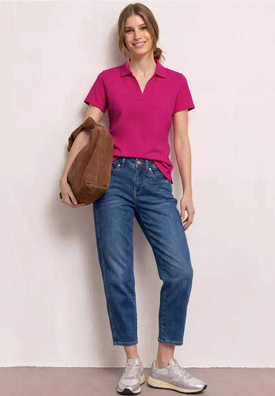 Een persoon in een roze polo, blauwe jeans, sneakers en bruine schoudertas voor een witte achtergrond.