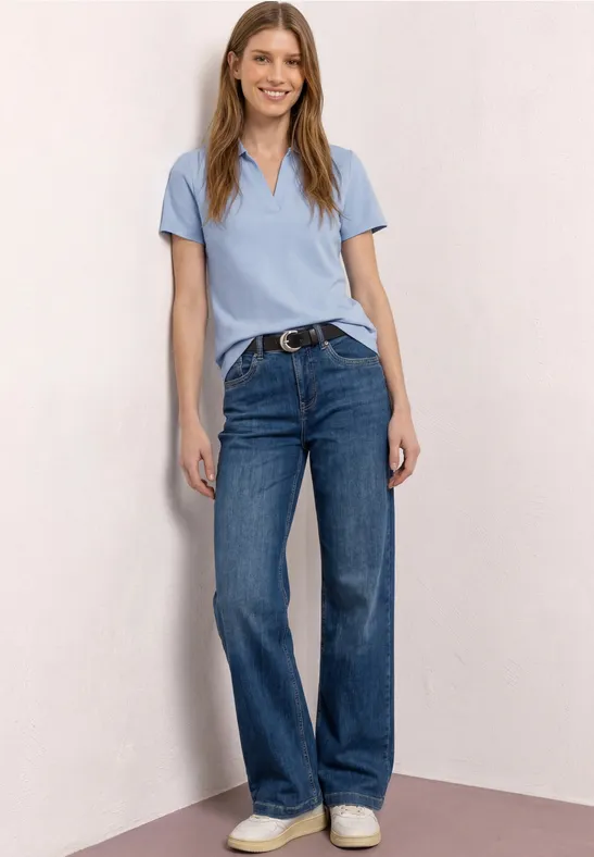 Een vrouw in lichtblauw poloshirt, blauwe jeans en witte sneakers staat nonchalant voor een neutrale muur.