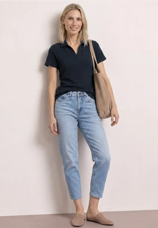 Vrouw staat voor een lichte achtergrond, draagt een zwart polohemd, blauwe jeans, bruine loafers en een schoudertas.