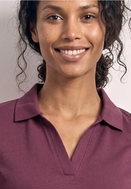 Een persoon met krullend haar draagt een paarse polo. Alleen het bovenlichaam en de hals zijn zichtbaar.