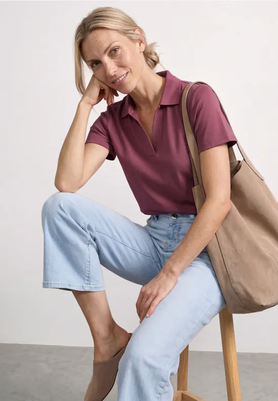 Eine Person sitzt auf einem Hocker, trägt lila Poloshirt, helle Jeans, bequeme Schuhe und eine Tasche.