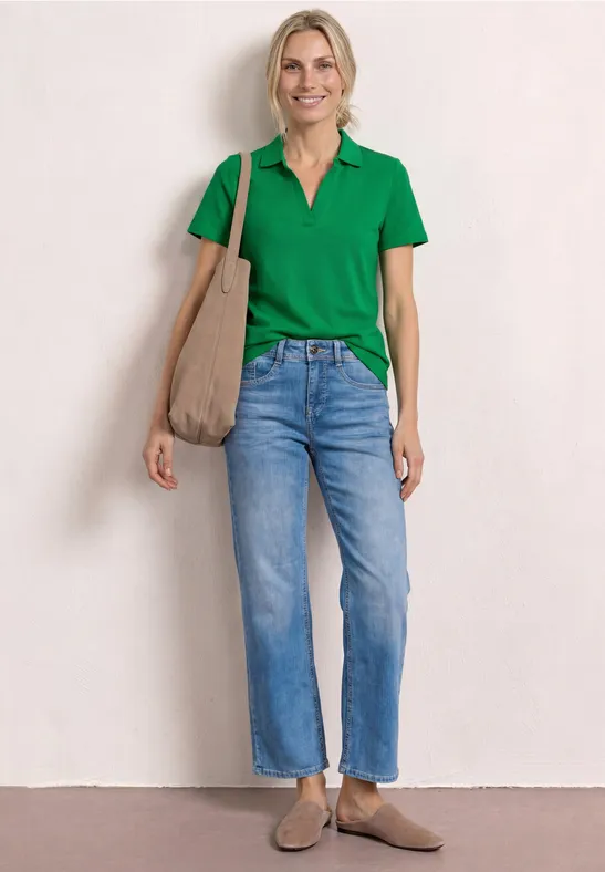 Frau in grüner Polo, Jeans, beigem Shopper und Mokassins vor heller Wand.