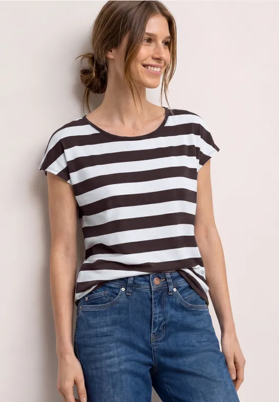 Eine Frau trägt ein gestreiftes T-Shirt und Jeans vor hellem Hintergrund.