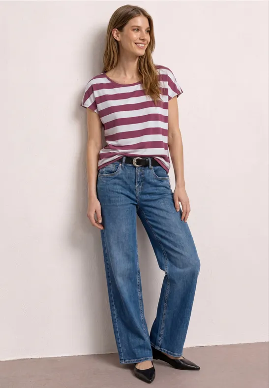 Vrouw met gestreept T-shirt, wijde spijkerbroek en platte zwarte schoenen, staand voor een lichte achtergrond.