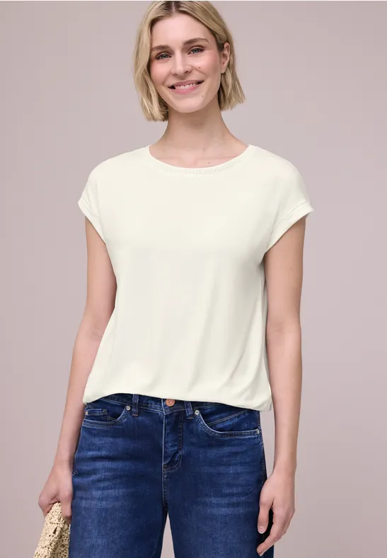 Frau trägt schlichtes weißes T-Shirt und blaue Jeans, steht vor neutralem Hintergrund.