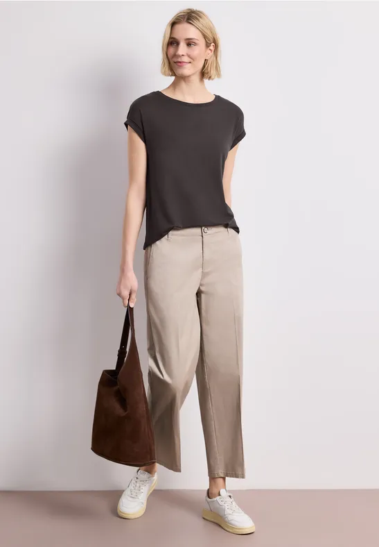 Een persoon draagt een zwart t-shirt, beige broek, witte sneakers en houdt een bruine tas vast.