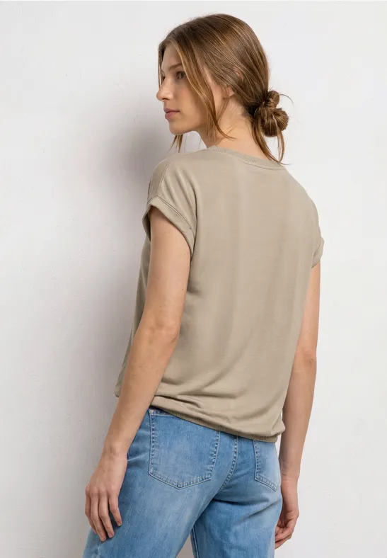 Frau in beigem T-Shirt und Jeans steht mit dem Rücken zur Kamera, die Haare zu einem niedrigen Dutt gebunden.