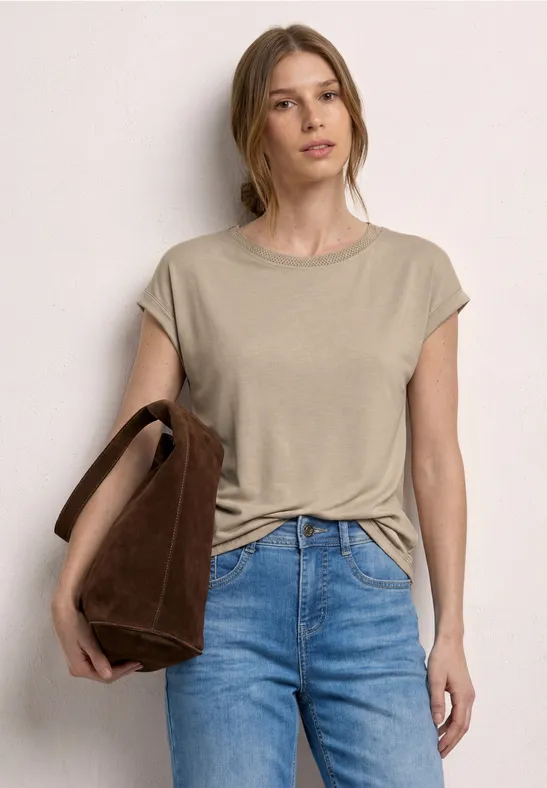 Frau in beige Kurzarmshirt, blauen Jeans trägt eine braune Schultertasche vor neutraler Wand.