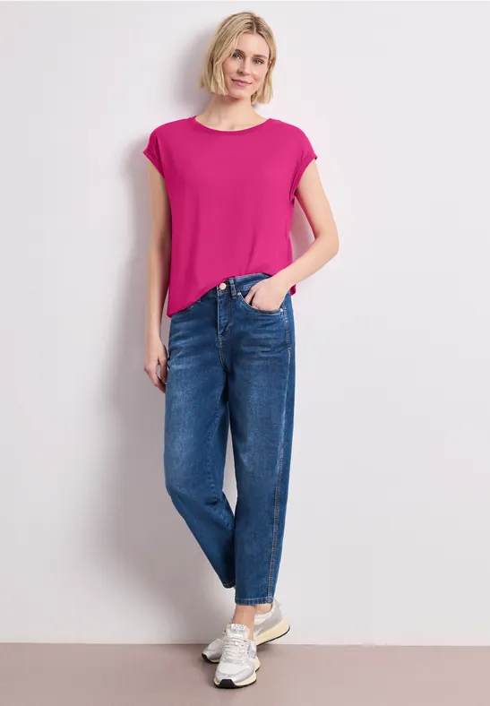 Frau in pinkem T-Shirt, legeren blauen Jeans und weißen Sneakern vor weißem Hintergrund.