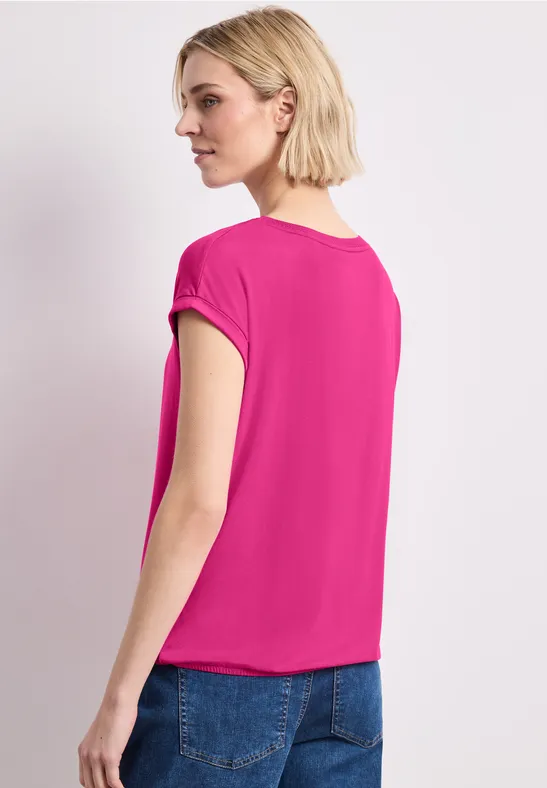 Frau von hinten in einem pinken Kurzarmshirt und blauen Jeans vor weißem Hintergrund.
