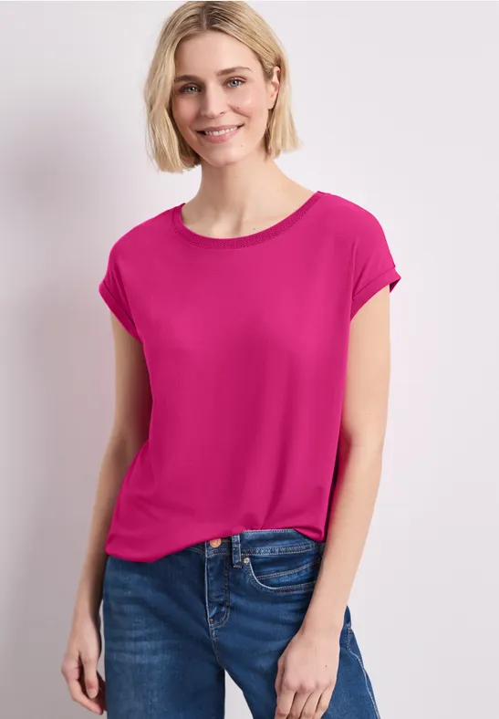 Modell trägt pinkes T-Shirt und blaue Jeans vor hellem Hintergrund.