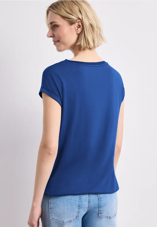 Persoon van achteren met blond haar, blauw T-shirt en lichtblauwe spijkerbroek tegen lichte achtergrond.