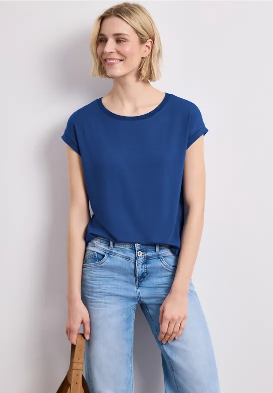 Vrouw in blauw T-shirt en lichte jeans voor een witte achtergrond, met handtas in linkerhand.