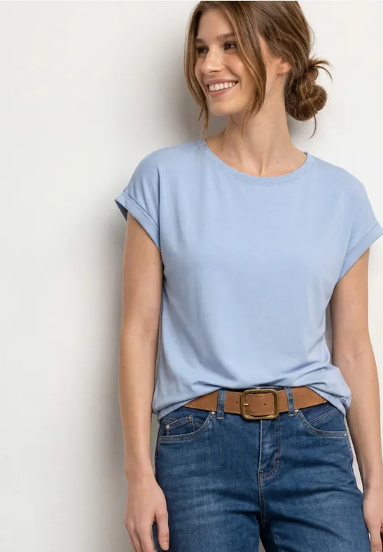 Eine Frau in hellblauem T-Shirt mit aufgekrempelten Ärmeln, Jeans und braunem Gürtel.