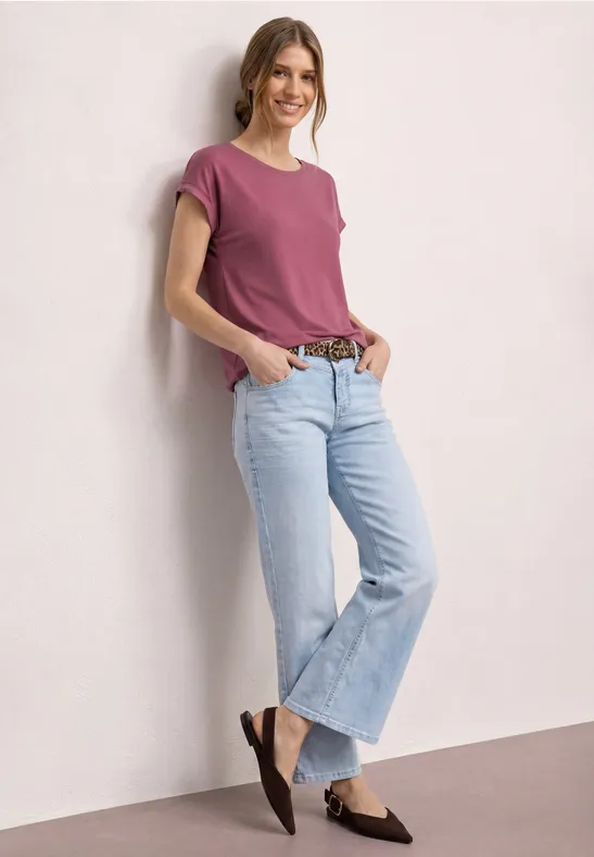 Eine Frau in pinkem T-Shirt, hellblauen Jeans und braunen Slippern lehnt lässig an einer hellen Wand.