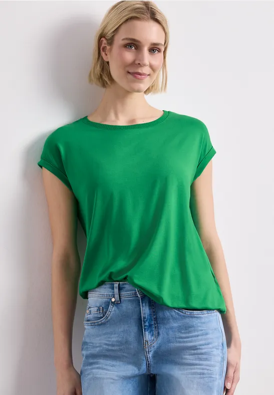 Frau in grünem T-Shirt und blauer Jeans vor weißem Hintergrund.