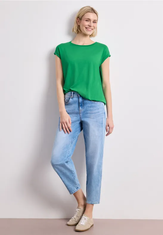 Ein Modell steht vor weißem Hintergrund in grünem T-Shirt, hellblauen Jeans und beigen Sneakern.