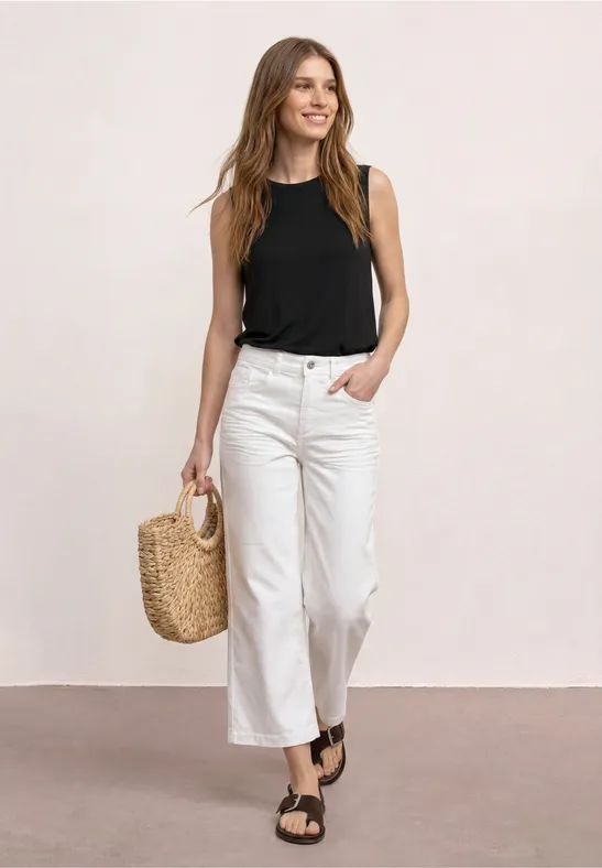 Vrouw in witte culotte, zwart tanktop, rieten tas en zwarte sandalen voor lichte achtergrond.