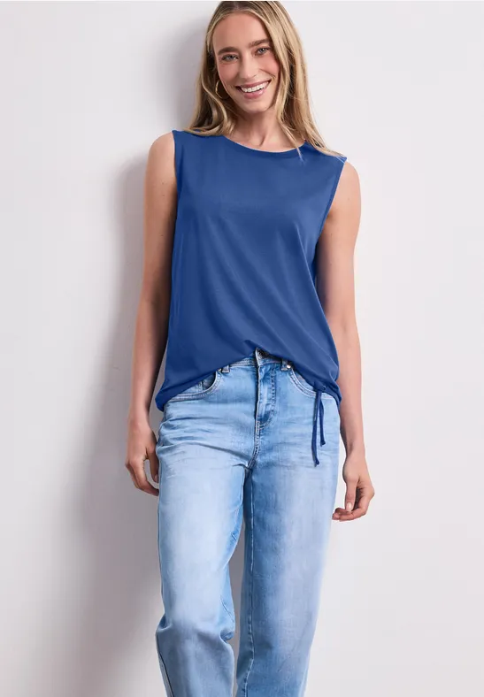 Eine Frau trägt ein ärmelloses blaues Oberteil und helle Jeans vor weißer Wand.