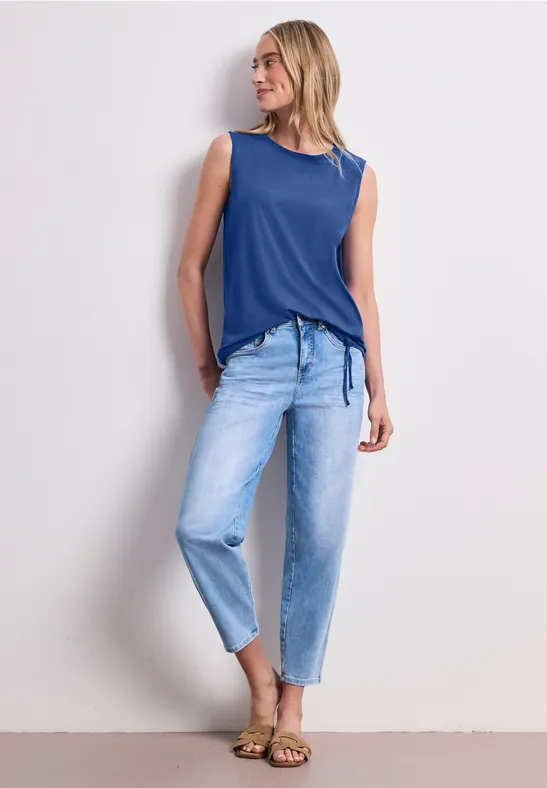 Eine Blonde posiert in ärmellosem blauem Top, hellen Jeans und Sandalen vor hellem Hintergrund.
