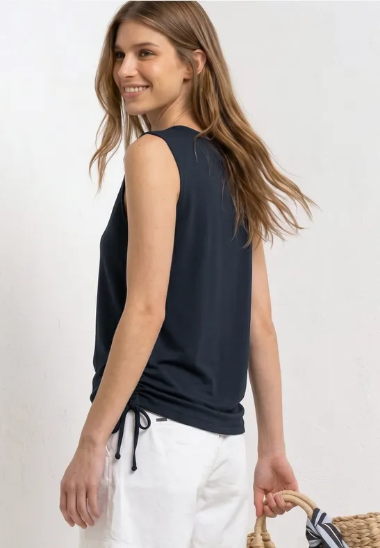 Vrouw in een zwarte mouwloze top, witte broek en strooien tas, ontspannen staand op de foto.