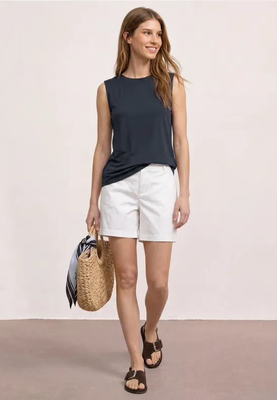 Vrouw in marineblauw topje en witte shorts, met een gevlochten tas en zwarte sandalen voor een neutrale achtergrond.