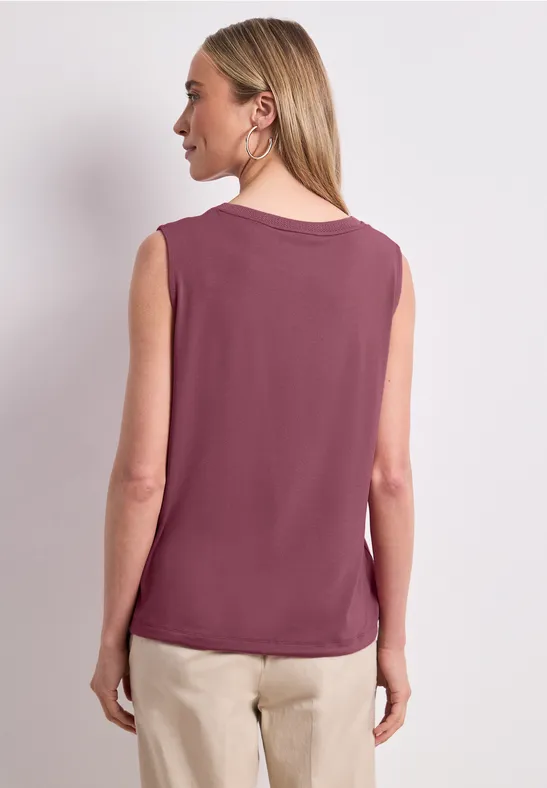 Eine Frau steht Rücken zur Kamera, trägt ein ärmelloses violettes Top und helle Hose.