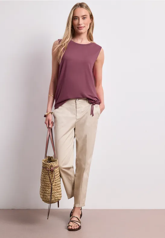 Frau trägt ärmelloses lila Top, helle Chinos, schwarze Sandalen und Korbtasche.
