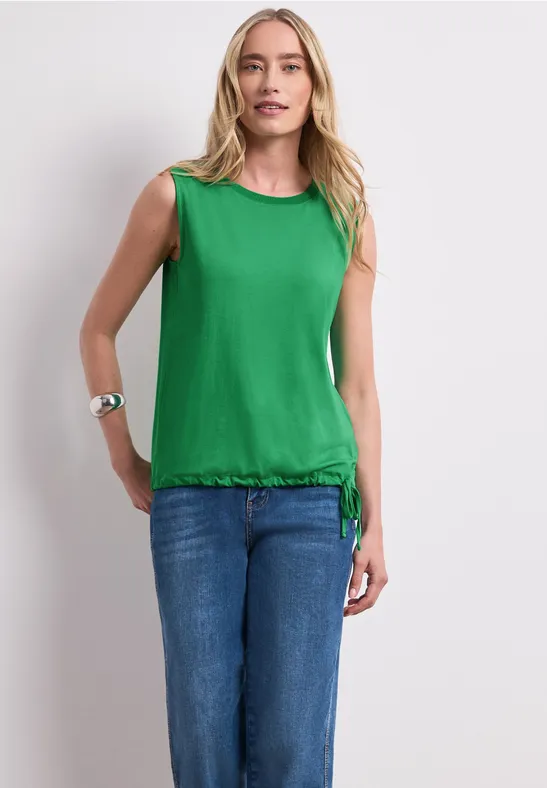 Eine Frau trägt ein grünes ärmelloses Top mit Kordelzug und blaue Jeans, steht vor weißem Hintergrund.
