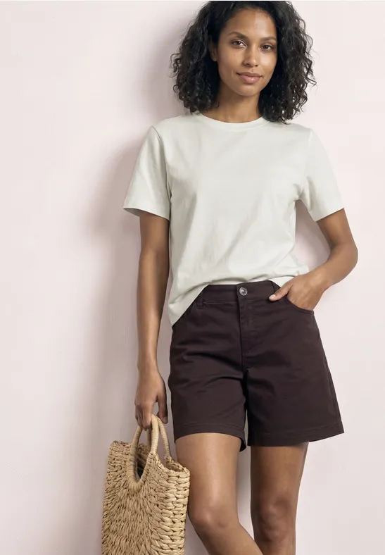 Vrouw in witte T-shirt en bruine short met gevlochten strooien tas, lichte achtergrond.