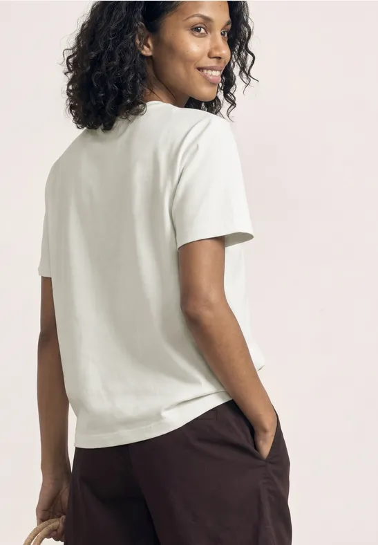 Rugaanzicht van een persoon met krullend haar in een wit shirt en een bruine broek.