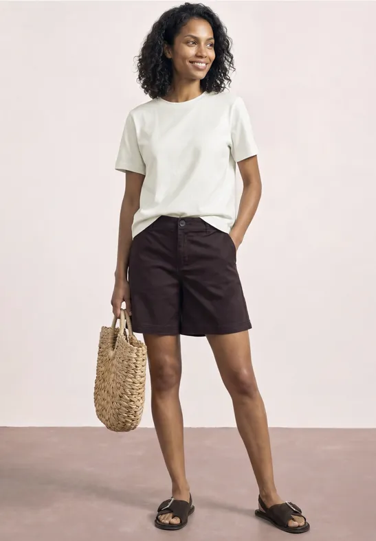 Vrouw in wit T-shirt, zwarte shorts en sandalen, met een rieten tas in haar hand.
