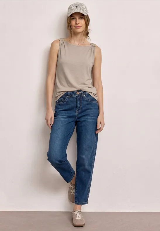 Een persoon staat voor een lichte muur met een beige tanktop, blauwe jeans en bijpassende loafers.