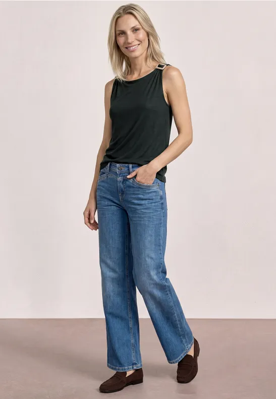 Vrouw in zwart mouwloos topje en blauwe jeans, ontspannen pose met handen in de zakken.