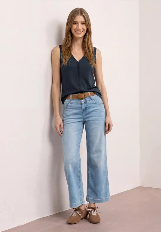 Een vrouw met een donkerblauw hemdje, lichte wijde jeans en bruine schoenen voor een lichte muur.