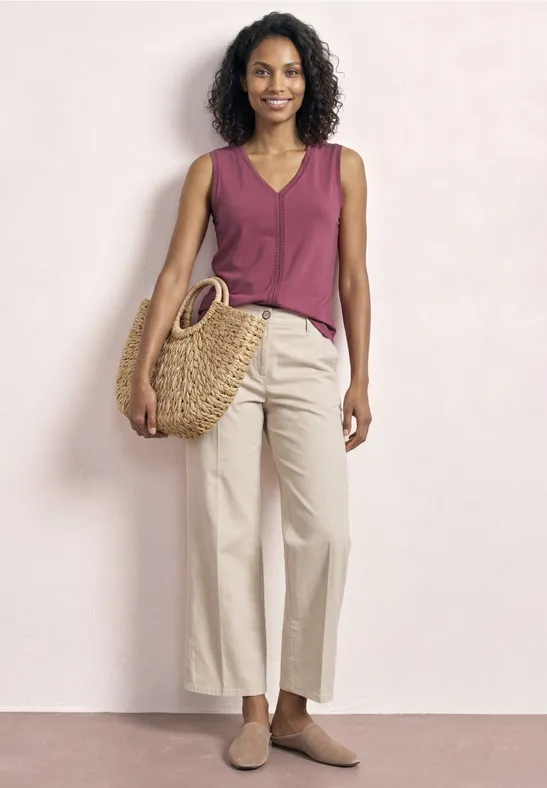 Vrouw met krullend haar in een bordeaux top, beige wijde broek, rieten tas en platte schoenen.