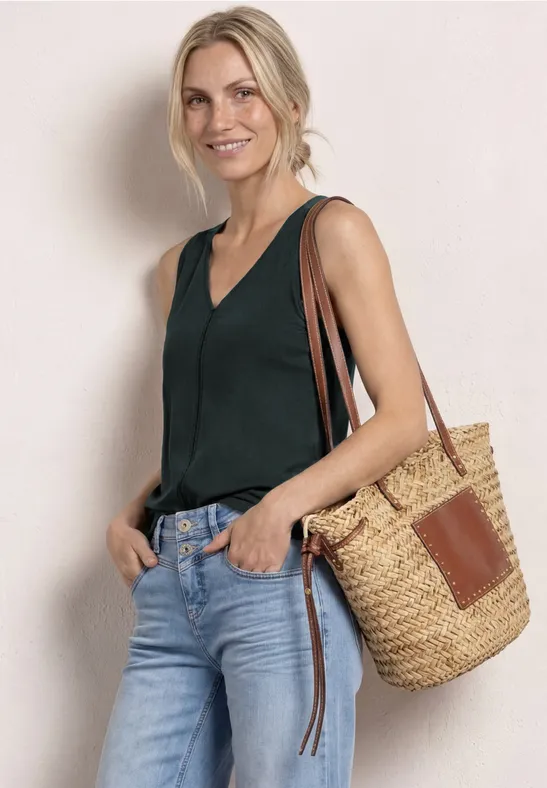Vrouw in groen mouwloos topje, jeans en strooien tas, nonchalant pose tegen lichte achtergrond.
